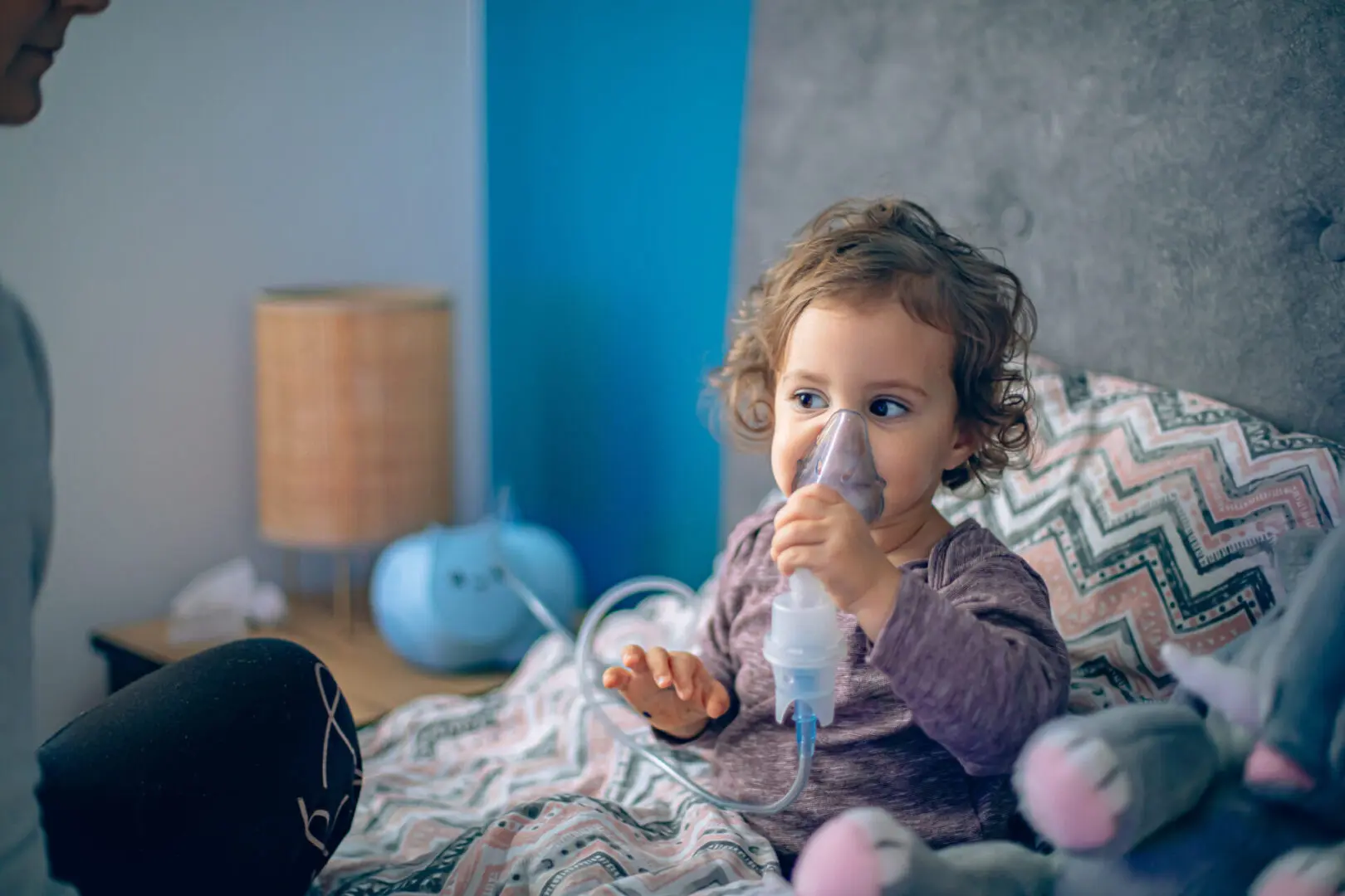 Brave little girl having inhalation in bed Sick baby girl holding inhalator with mother next to her